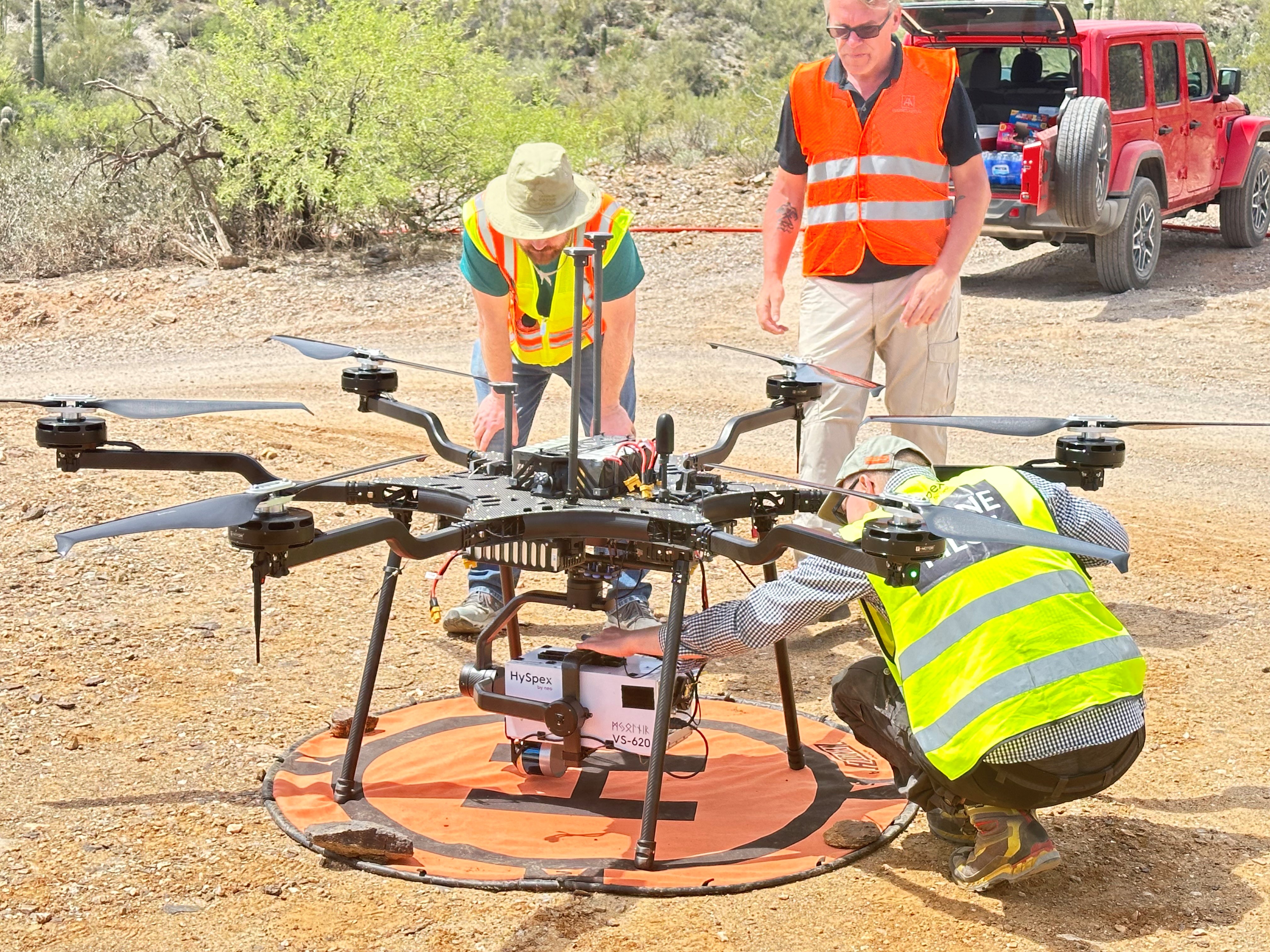 Three men in saftey vests and hard hats stand around a large drone
