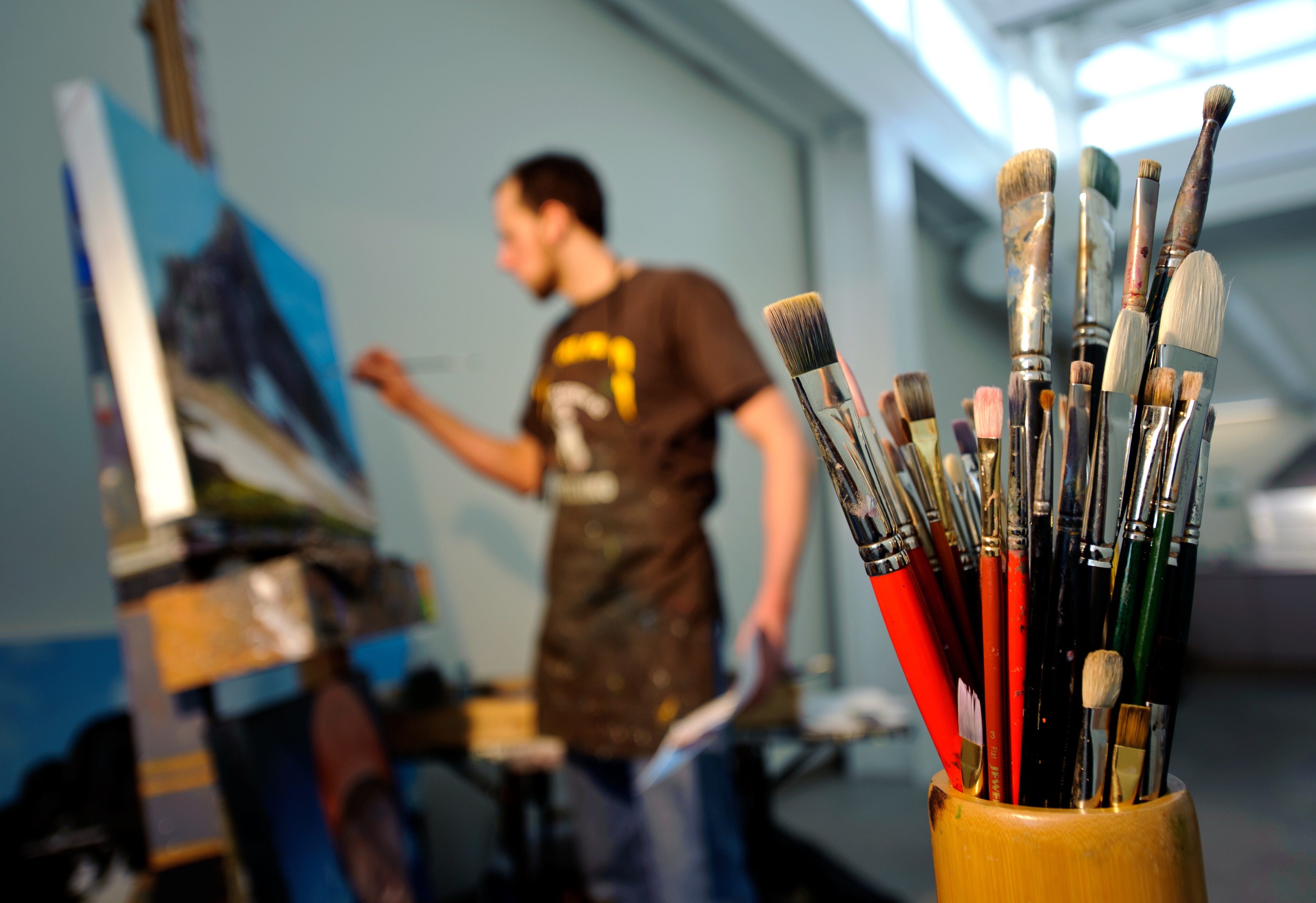 student in studio working in background with cup with paintbrushes in it in forground
