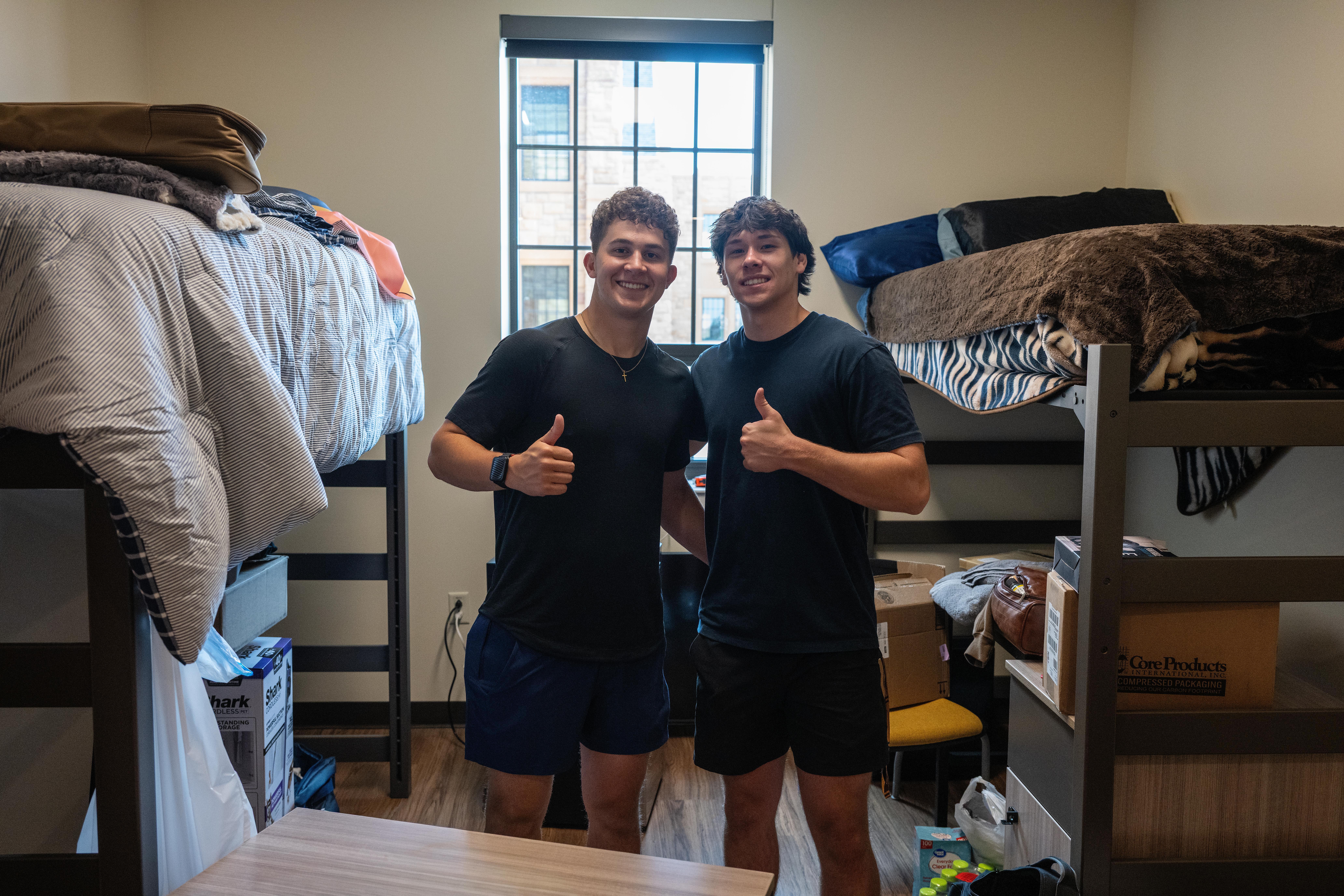 Two men holding thumbs up in a dorm room