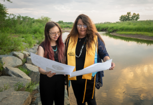 2 women standing by a river looking at papers
