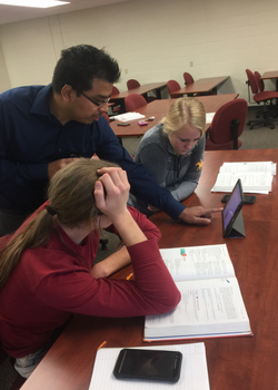 two female students with books open working on an ipad with the professor standing in between