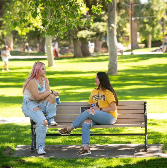 Two students sitting on a bench and talking in the green area along Ivinson Street.