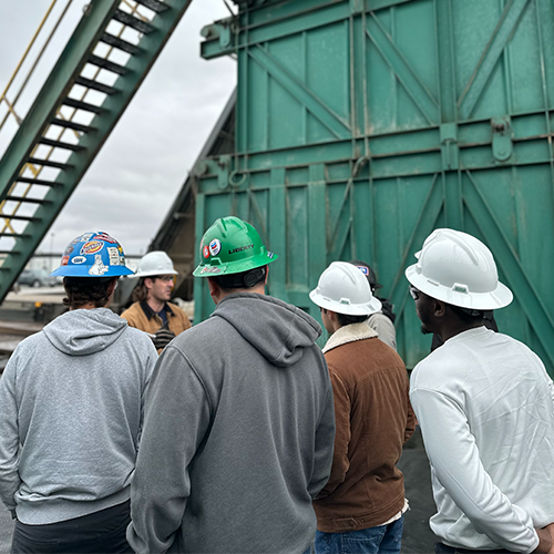 Students wearing hard hats looking at an oil rig in the field