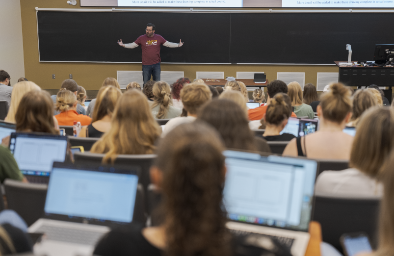 A professor speaking at the front of a large classroom of students.