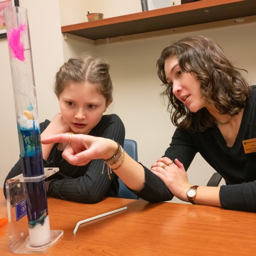 A woman and a child looks at a tall beaker with blue liquid inside.