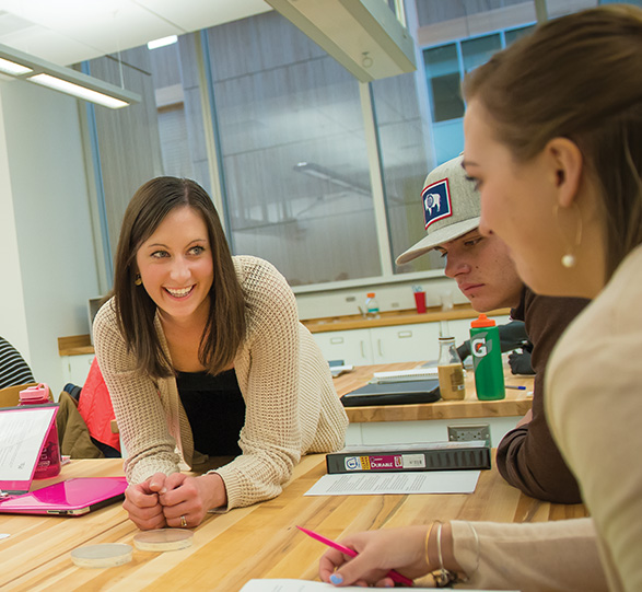 Teacher leaning on table talking to students. 