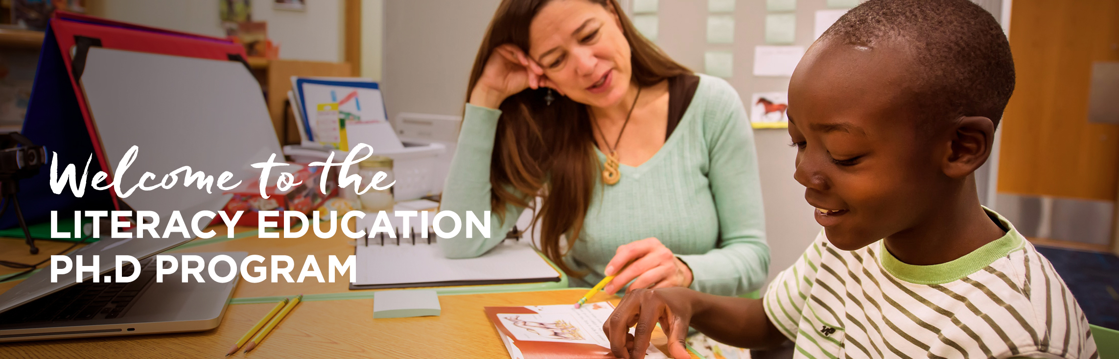 University of Wyoming Literacy Education Ph.D. Student works with a young student who is reading a book at a table. 