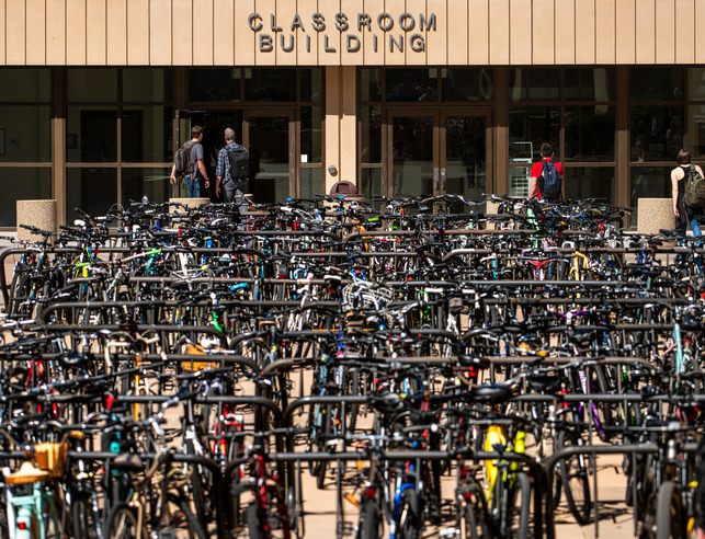 A large amount of bicycles in the bike rack with the classroom building in the background