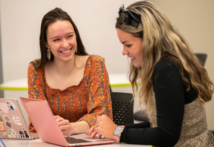 A UW staff member and student smile at each other as they both work at laptops in a UW help center on campus.