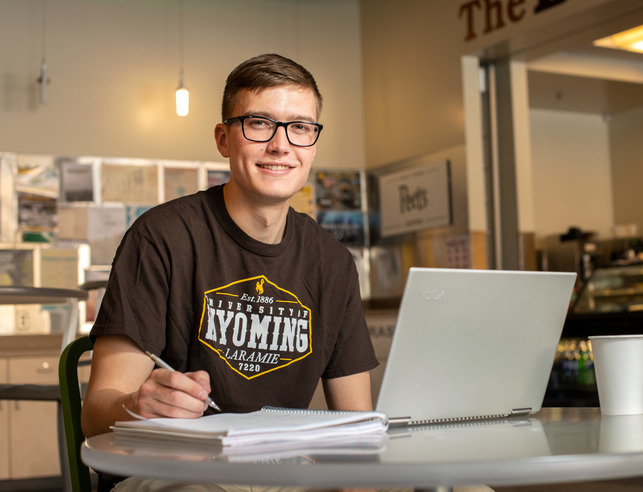A student in a coffee shop doing work on their computer and notebook