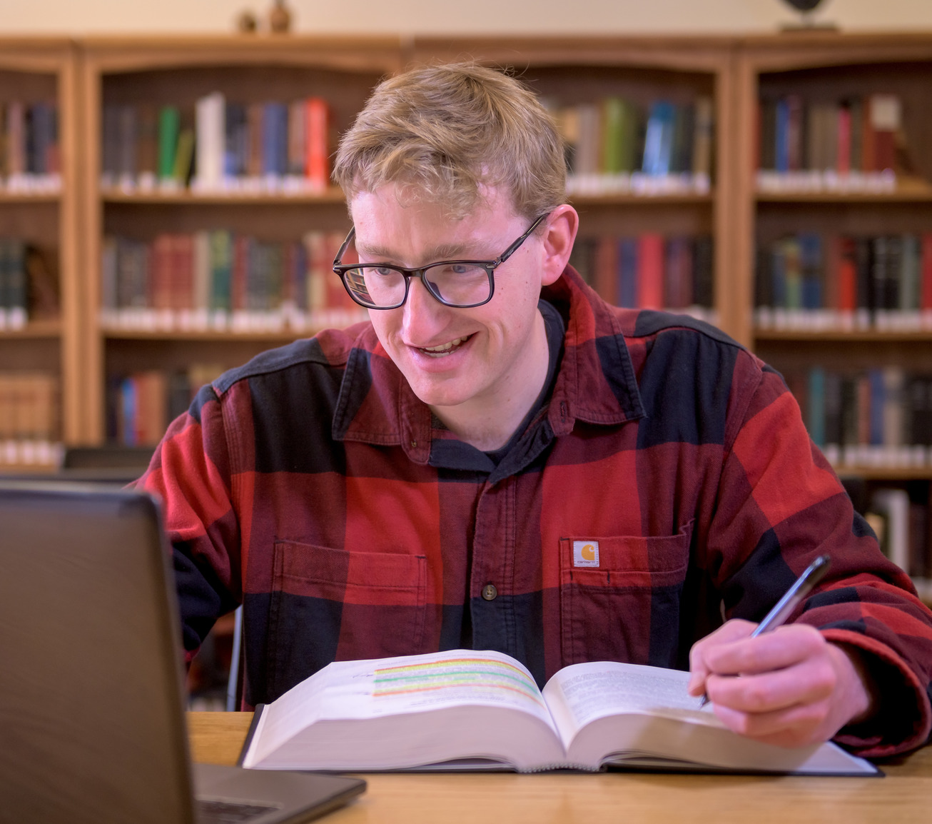 A student in a red and black plaid shirt studies with books in the background.