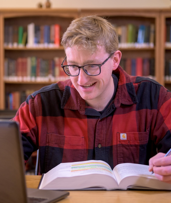 A student in a red and black plaid shirt studies with books in the background