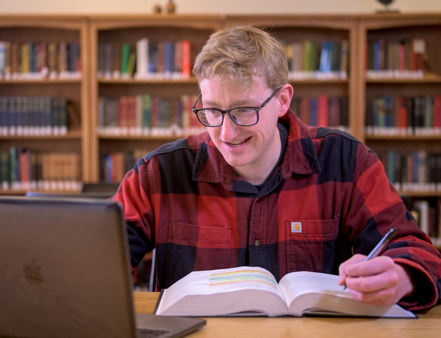 A student in a red and black plaid shirt studies with books in the background.
