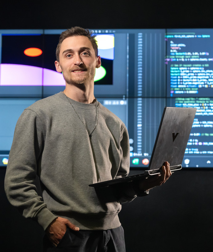 A student smiles for a photo while holding a laptop with code in the background