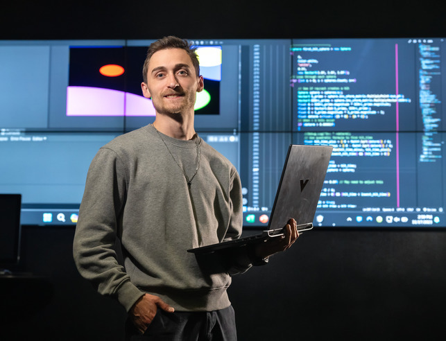 A student smiles for a photo while holding a laptop with code in the background