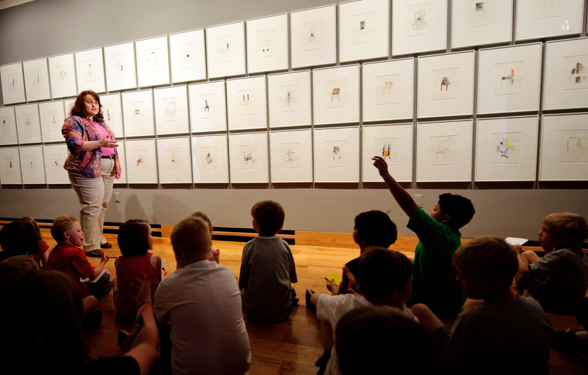 a teacher is standing in front of several art works and speaking to a group of children