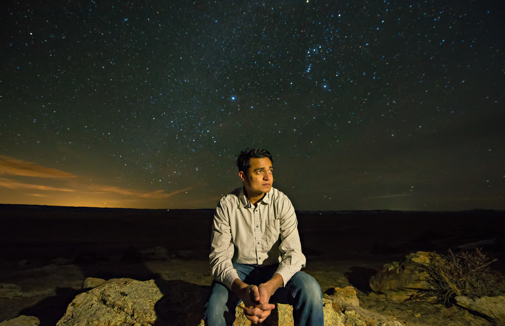 A person sits on a rock staring off to the side in front of the night sky with a lot of bright stars