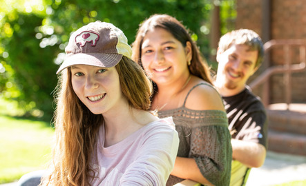 Three students smile for a photo while sitting on a bench