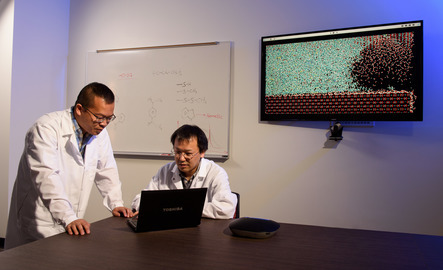 Two chemists wearing lab coats are using a computer and behind them is a white board with chemistry symbols written on it and a monitor screen with data visualization