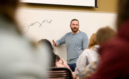 Students and instructor in a classroom where the whiteboard has chemistry symbols written on it