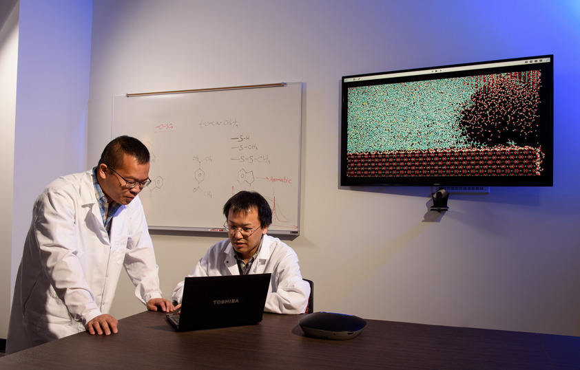two Asian men wearing lab coats are using a computer and behind them is a white board with chemistry symbols written on it and a monitor screen with data visualization