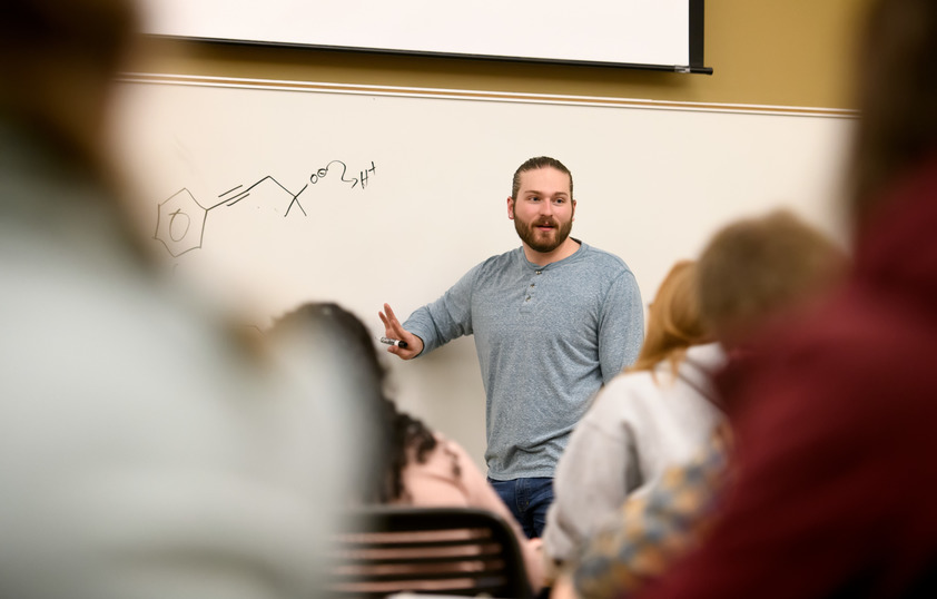 students and instructor in a classroom where the whiteboard has chemistry symbols written on it