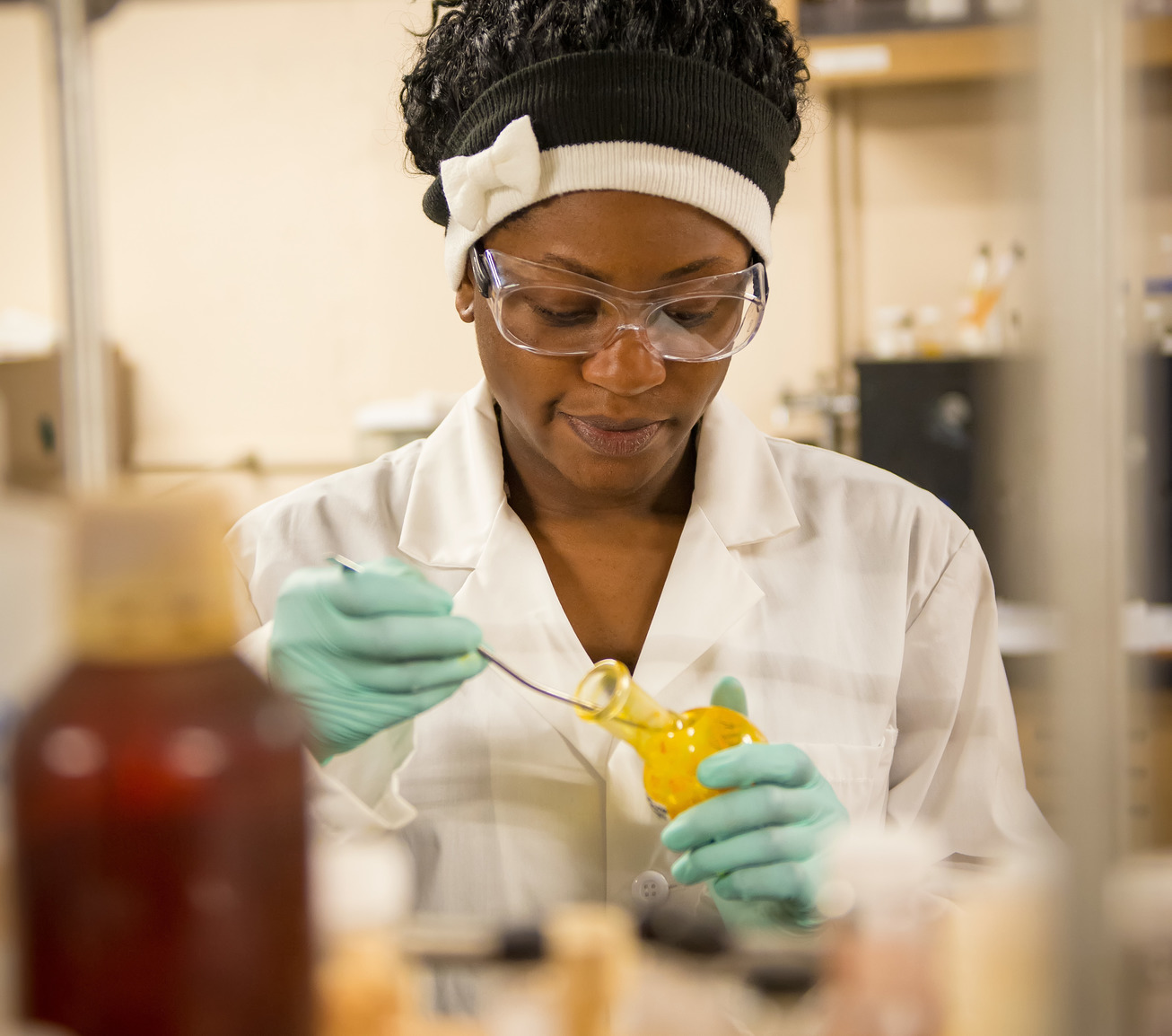 a young Black woman wearing a lab coat, gloves, and safety goggles, is using the equipment in a chemistry lab