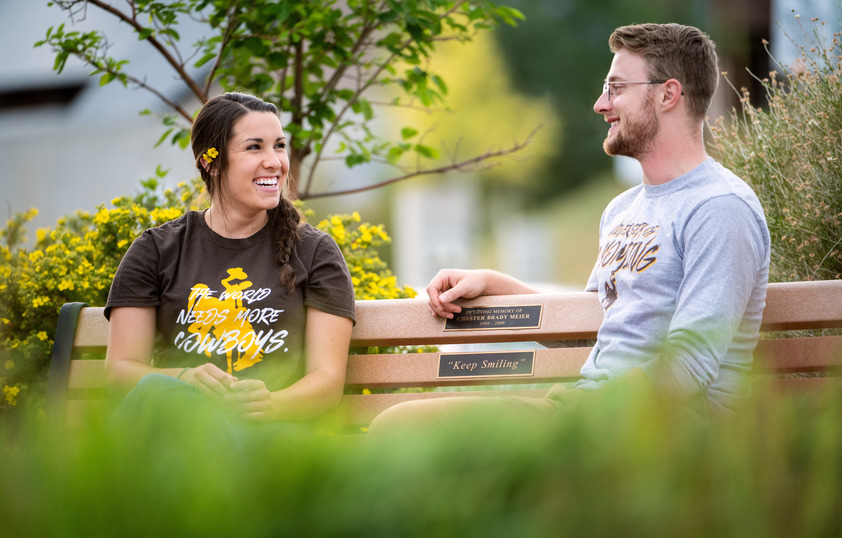 Two students sit on a bench next to green trees