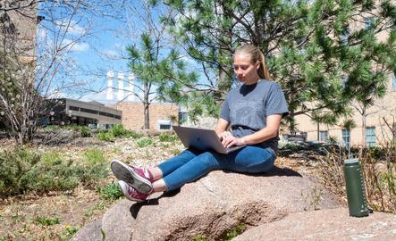 A student sits on a rock while working on their laptop
