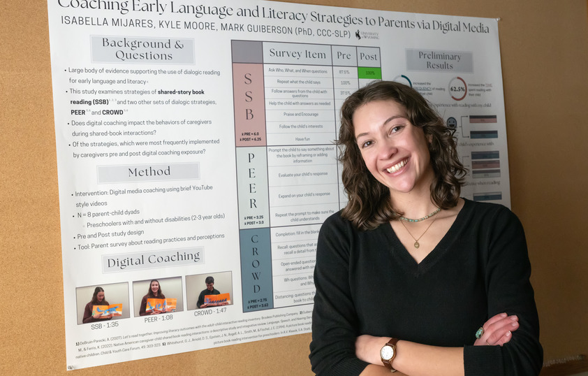 A graduate student poses for a picture in front of a speech langugae pathology poster