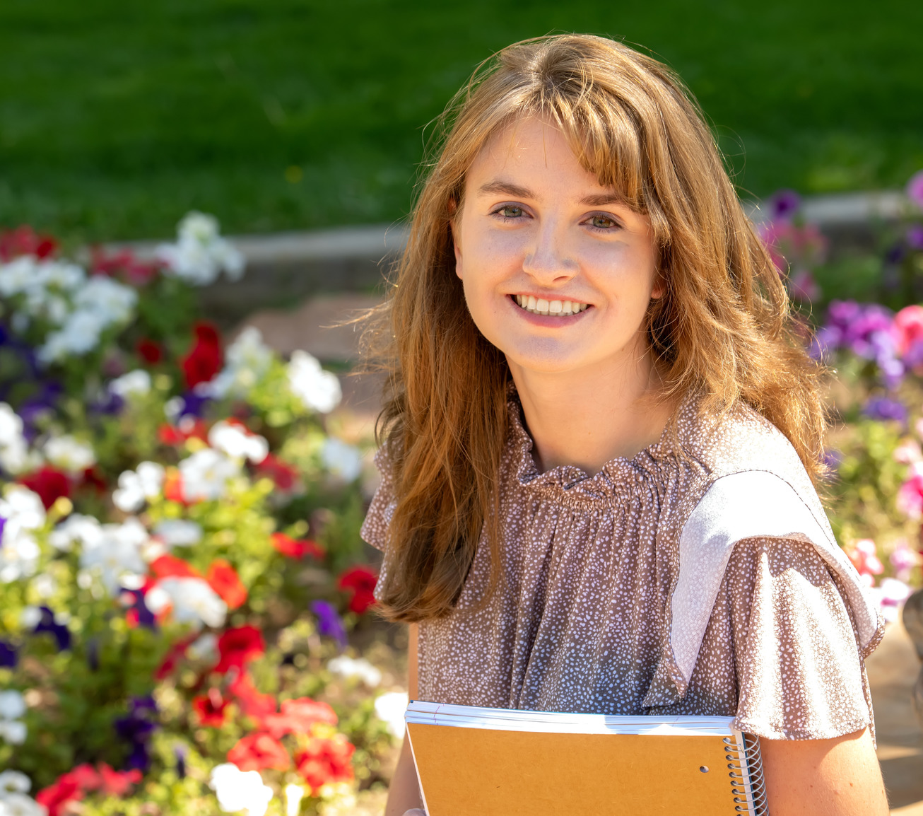 Student smiling and posing for a photo in front of flowers with a notebook in their hand