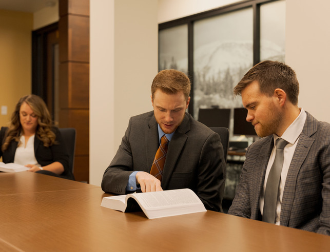 Two people look at a book together in a conference room with one person looking at a book alone in the background