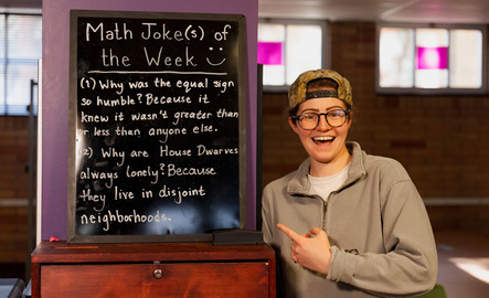 A math teacher poses with a chalk board displaying math jokes