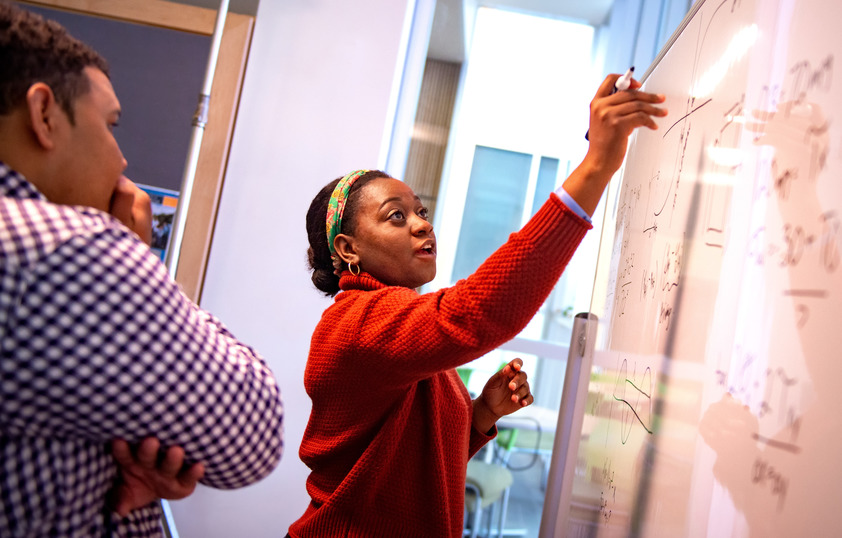 A student writes math equations on a whiteboard while another student studies it