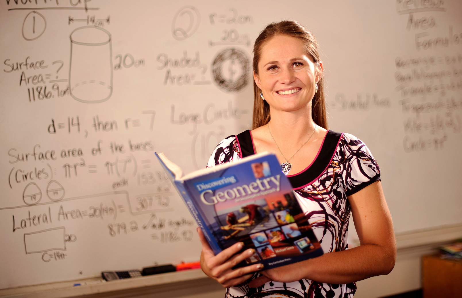 A math teacher smiles for a photo in her classroom while holding a geometry book