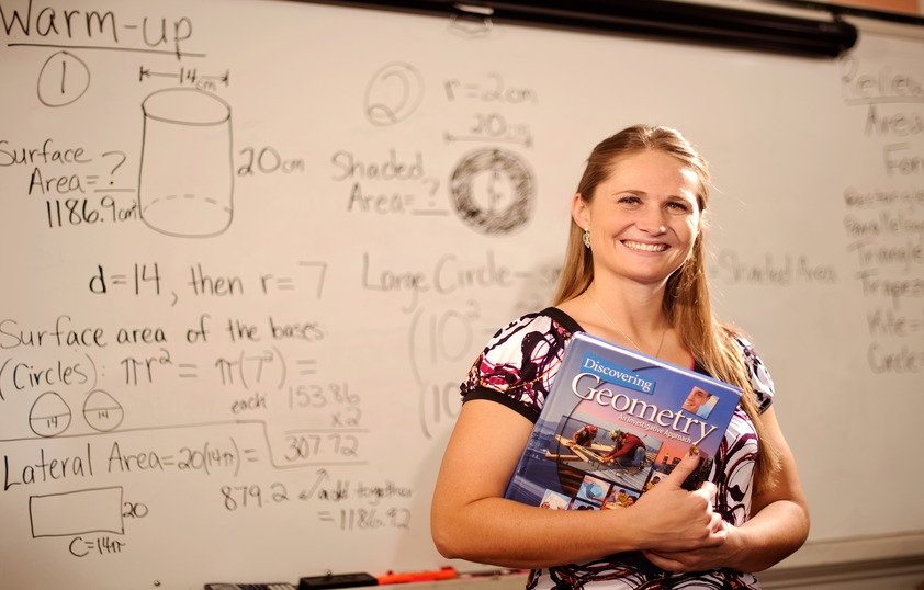 a smiling woman is holding a geometry book and standing in front of a whiteboard with geometry equations written on it