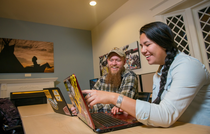 Two smiling students are using laptop computers while sitting at a shared table