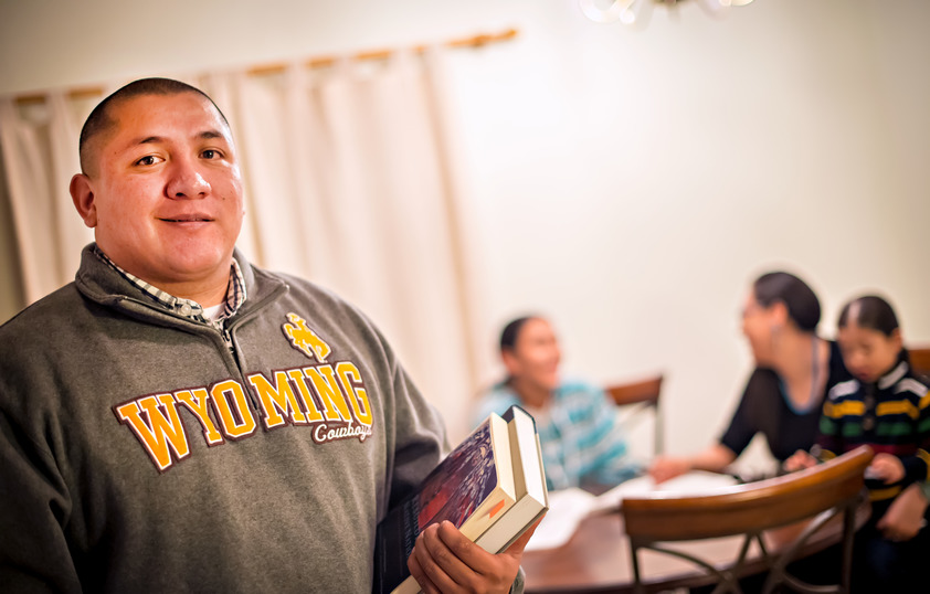 A University of Wyoming student is holding books in the foreground with some other people are sitting at a table in the background