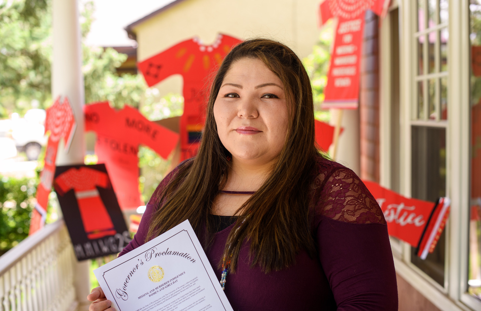 Taryn Jim, of the Northern Arapaho tribe, and recent Native American and Indigenous Studies graduate, is standing outside the Native American Education, Research and Cultural Center holding a Governor's Proclamation document and there are placards in the background spreading awareness about missing and murdered indigenous women.