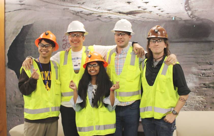 A group of students smile for a photo with hard hats and safety vests on