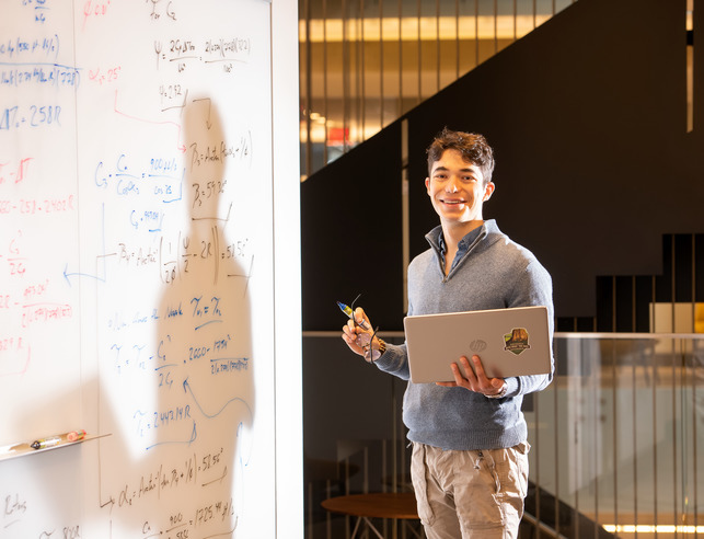 Online instructor holding laptop near whiteboard