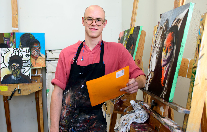 A student poses for a picture next to their painting on a canvas