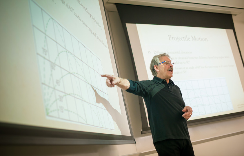 A professor stands at the front of the class teaching while pointing at projection screen