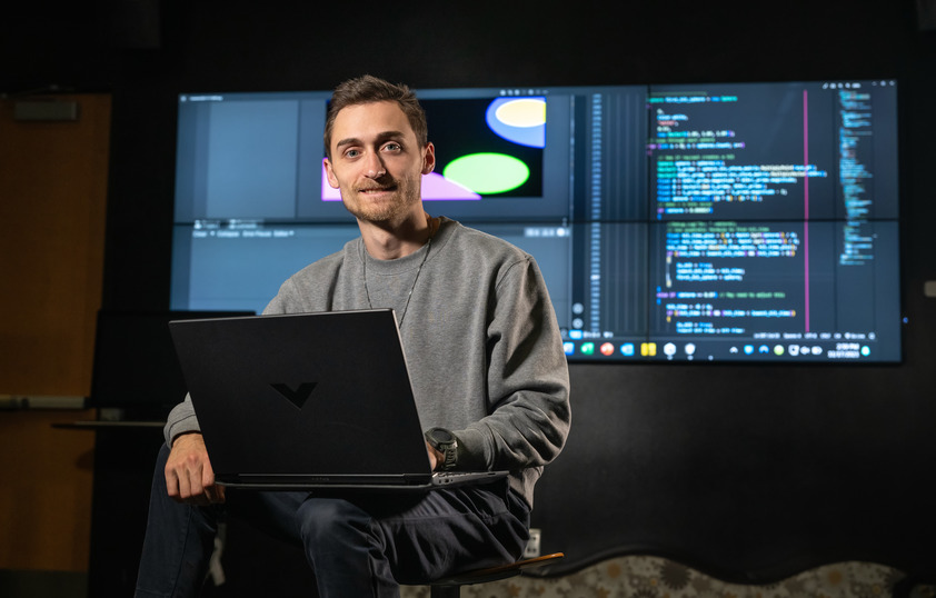 A student smiles for the camera with his laptop propped on his lap and code featured behind him