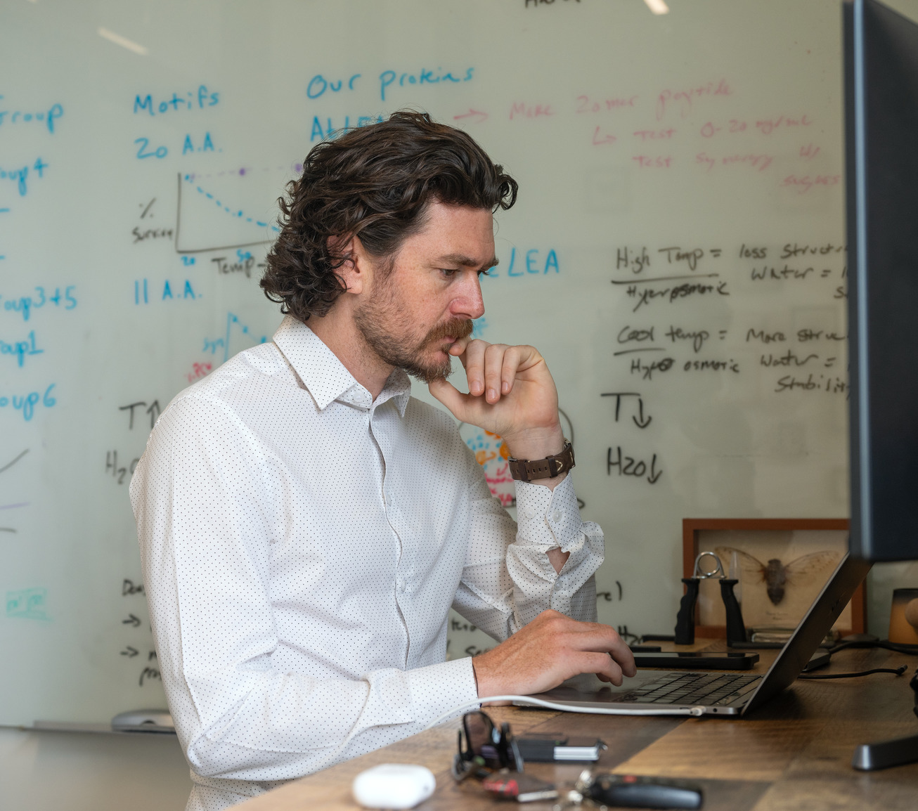 A professor looks at his laptop in his office with mathematics formulas on a whiteboard behind him