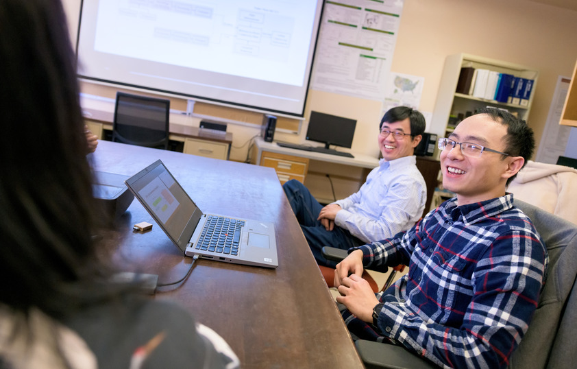 Three people sit at a conference table together laughing with their laptops in front of them on the table