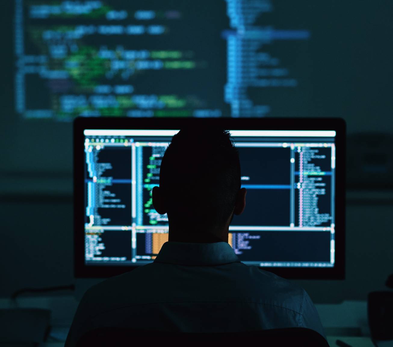 Silhouette of a person works on code in a dark room on a computer screen and a projector screen in the background