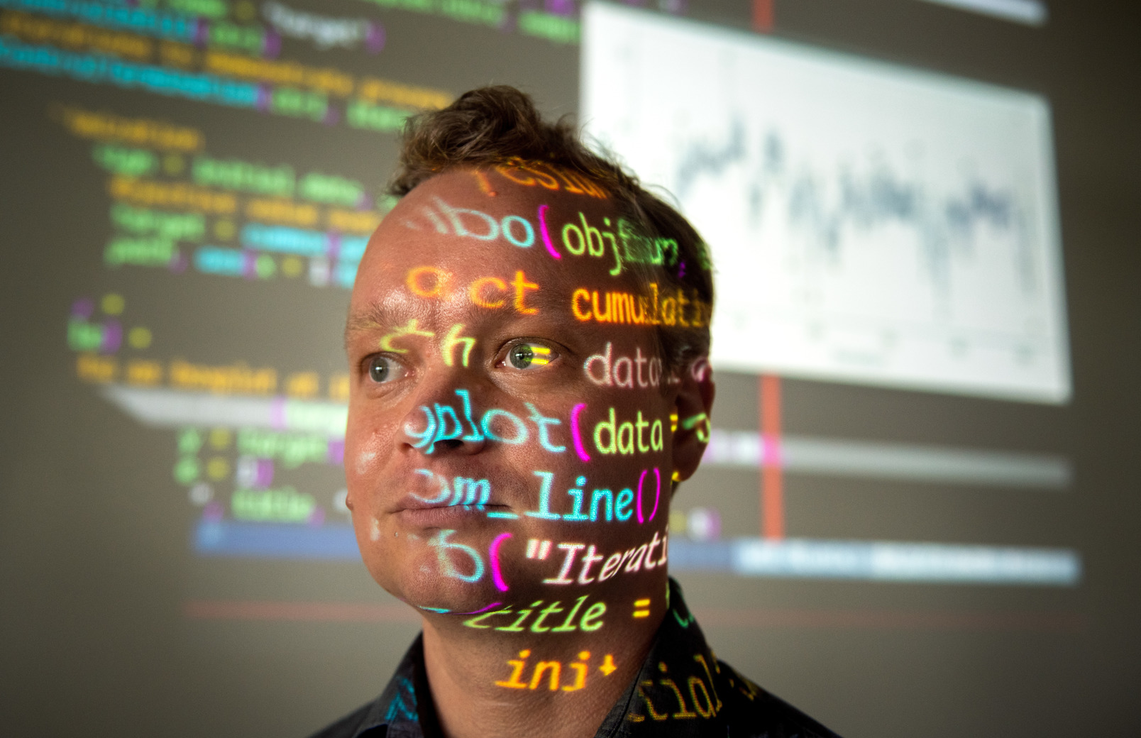 A person stands in front of a projector screen displaying code on his face