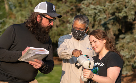 A professor helps two students with a surveying device