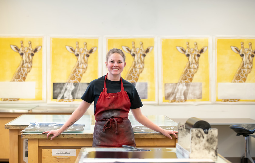 a smiling student in the printmaking studio standing in front of a series of prints